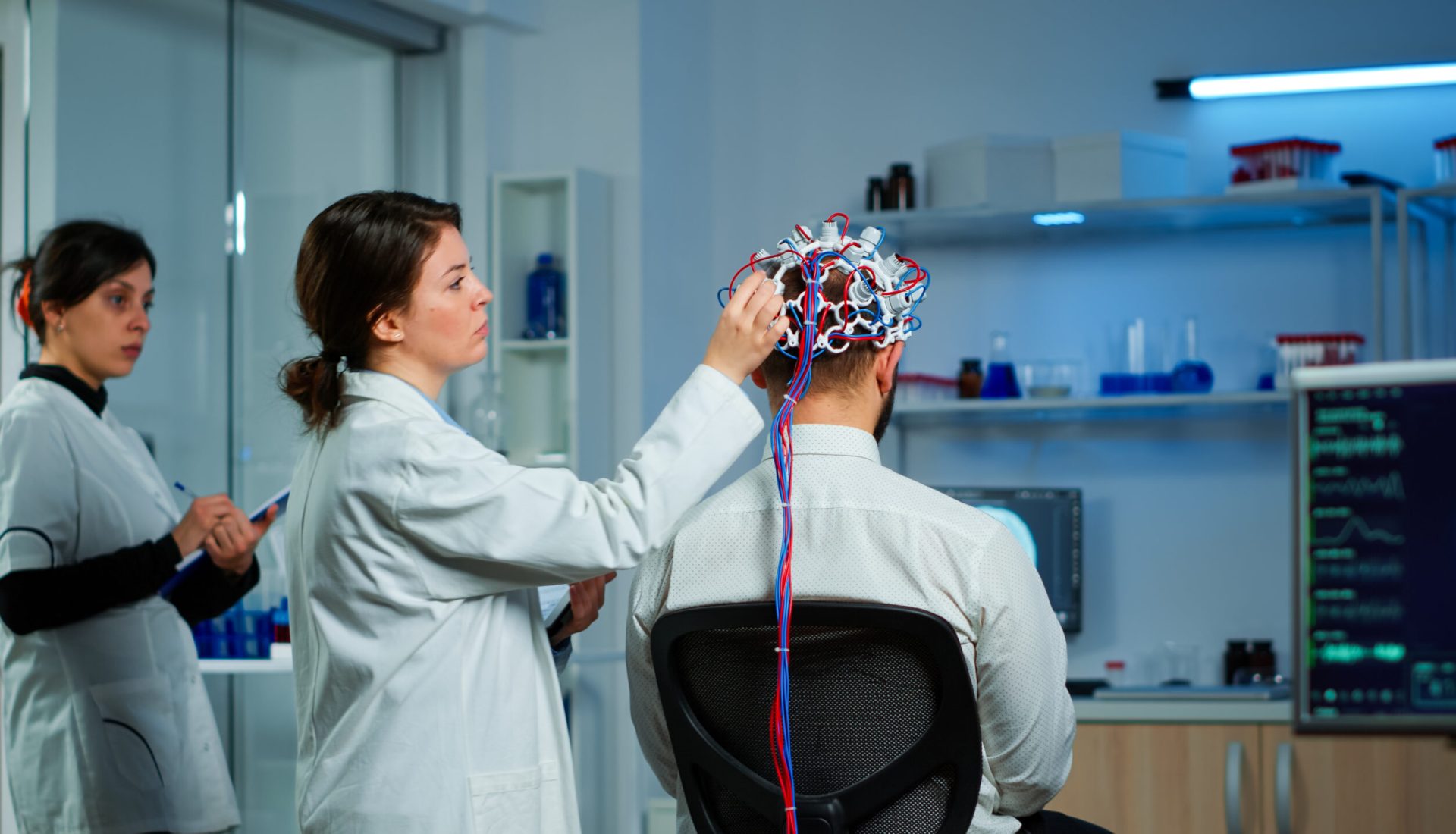 Back view of man patient wearing performant brainwave scanning headset sitting in neurological research laboratory while medical researcher adjusting it, examining nervous system typing on tablet.