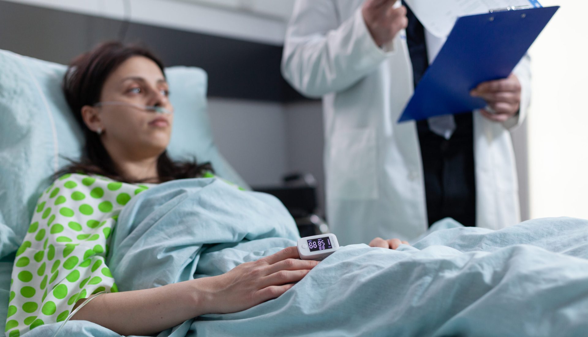 Woman on hospital bed with pulse oximeter on finger showing low oxygen saturation listening to doctor reading lab results from clipboard. Patient with nasal cannula recieving diagnosis results.